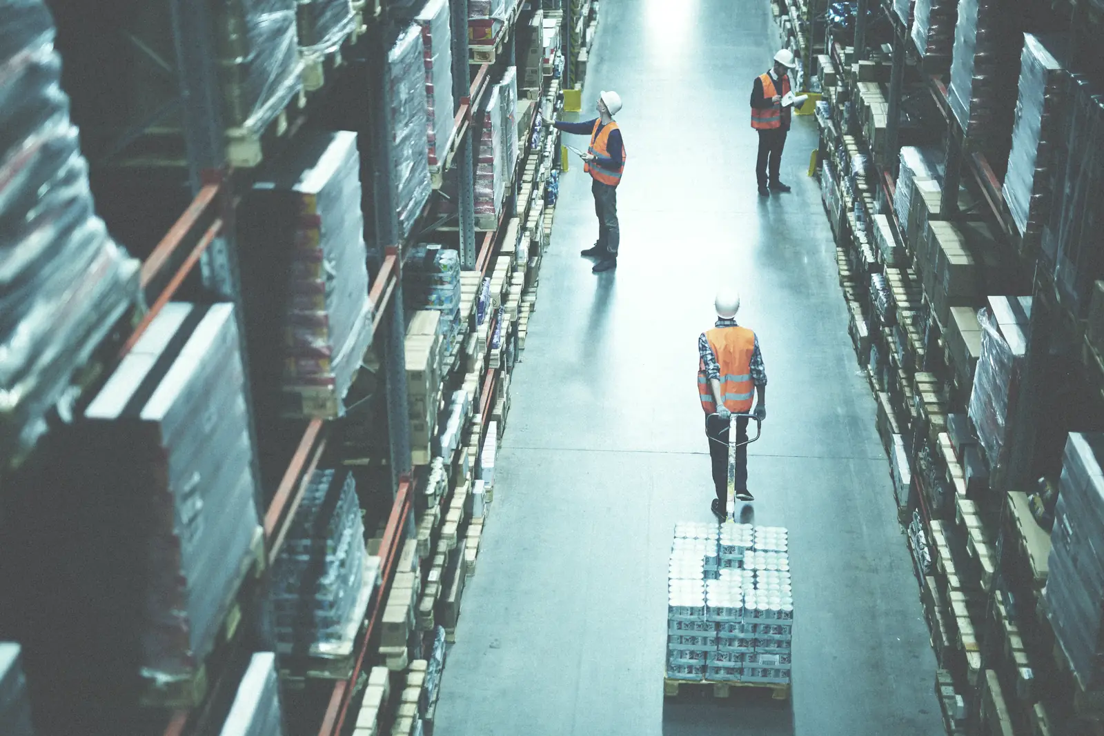 View down a warehouse aisle lined with high pallet racks, two workers in hi-vis vests walking between the rows