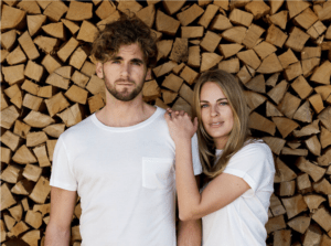 Das Bild zeigt zwei Models, einen Mann und eine Frau, die jeweils das MUNTAGNARD T-Shirt in weiß tragen, das erste plastikfreie T-Shirt vor einer Holzwand